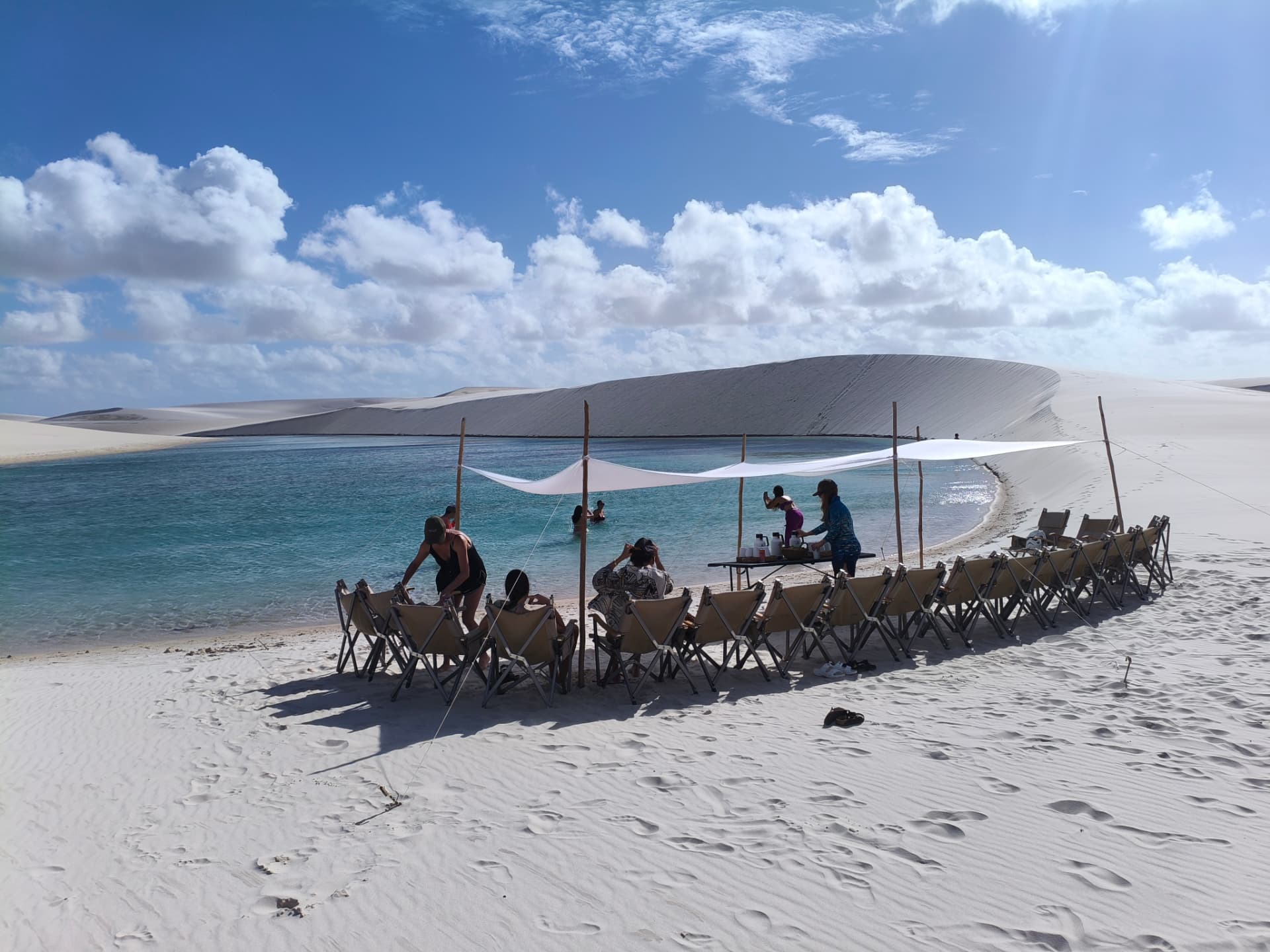 Visitantes em cadeiras sob tenda branca à beira de uma lagoa cercada por dunas em Santo Amaro do Maranhão