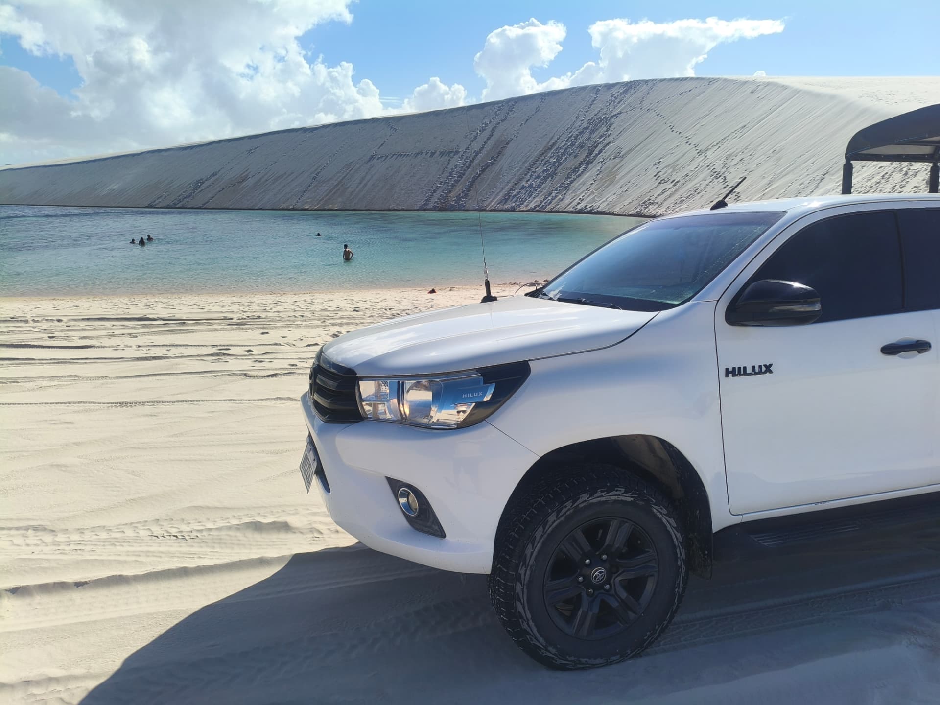 Veículo 4x4 branco na areia próximo a lagoa com banhistas e duna ao fundo