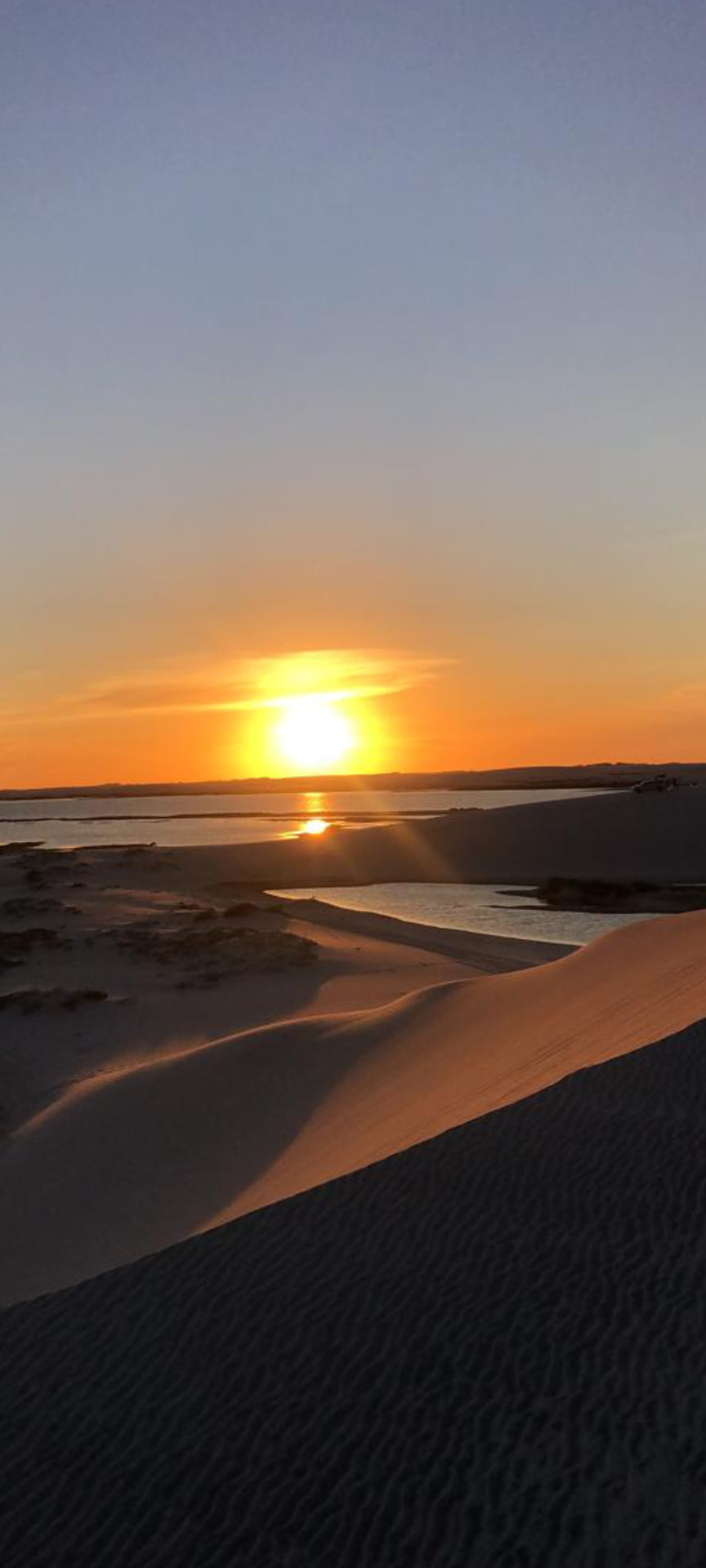 Pôr do sol alaranjado visto do alto das dunas com lagoa ao fundo
